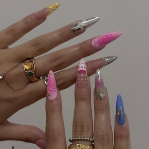 May include: Long almond-shaped artificial nails featuring diverse designs including metallic silver stars, pink ombre gradients, white polka dots, and raised textures. The nails are adorned with small gold studs and clear droplets, complemented by gold-toned rings.