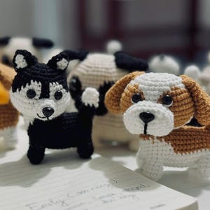 May include: A collection of handmade crochet dog figurines. The dogs are in various colors, including black and white, brown and white, and tan. Each dog has black button eyes and a black nose. The dogs are sitting on a white surface.