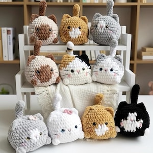 Può includere: Una collezione di peluche a forma di gatto fatti a mano all'uncinetto in vari colori, tra cui grigio, marrone, bianco e nero. Ogni gatto ha un corpo rotondo, una coda e un simpatico volto con dettagli ricamati. I peluche sono esposti su una mensola bianca.