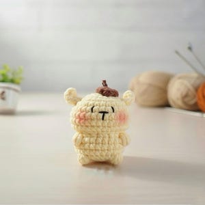 May include: A small, handmade crochet figure in a light yellow color, featuring a brown top and pink cheeks. The figure has a black 'T' shaped mouth and small arms and ears. The background includes yarn and knitting needles.