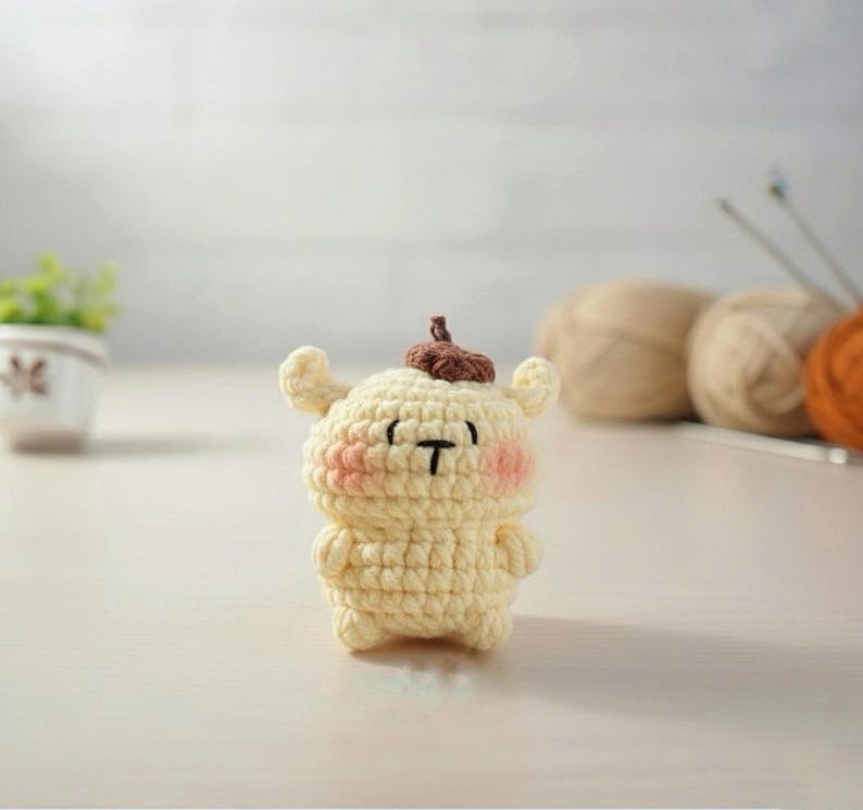 May include: A small, handmade crochet figure in a light yellow color, featuring a brown top and pink cheeks. The figure has a black 'T' shaped mouth and small arms and ears. The background includes yarn and knitting needles.