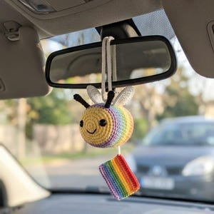 May include: A handmade crochet bee car charm with a rainbow body and a small rainbow flag attached. The bee has black antennae and a smiling face. The charm hangs from the rearview mirror in a car.