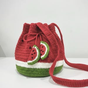 Puede incluir: Un bolso de cubo rojo tejido a ganchillo con un diseño de sandía. El bolso tiene un cuerpo rojo, una franja blanca y una base verde. Dos rodajas de sandía tejidas a ganchillo están unidas en la parte delantera. Tiene un cierre de cordón y una correa larga.