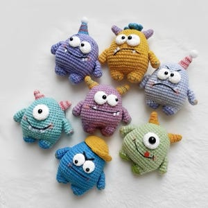 May include: A collection of seven colorful, handmade crochet monster toys. Each monster has a unique color, including blue, purple, yellow, and green. They feature large eyes, small horns, and some wear hats. The toys are arranged on a white surface.