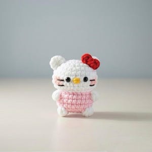 May include: A handmade crochet doll featuring a white head with a red bow, black eyes, and a yellow beak. The doll has a pink body and white arms and legs. The doll is standing on a white surface.