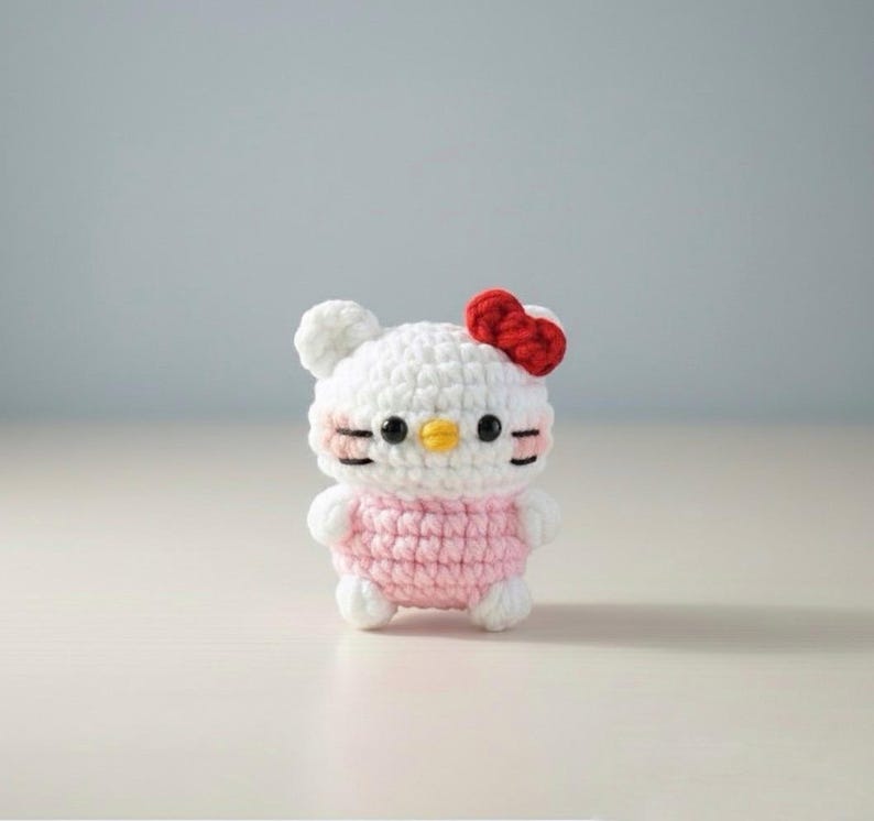 May include: A handmade crochet doll featuring a white head with a red bow, black eyes, and a yellow beak. The doll has a pink body and white arms and legs. The doll is standing on a white surface.