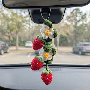 May include: A handmade crochet car charm featuring three red strawberries, white flowers with yellow centers, and green leaves. The charm hangs from a silver wire, adding a touch of whimsy to any vehicle's interior.