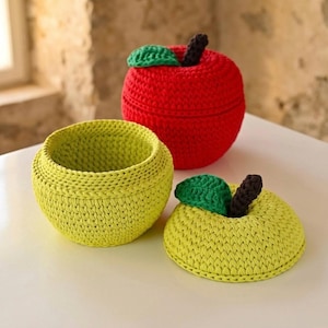 May include: Three handmade crochet apple-shaped items. One is red and lidded, another is open and yellow, and the third is a yellow apple-shaped lid. Each has a green leaf and a brown stem. The items are on a white surface.