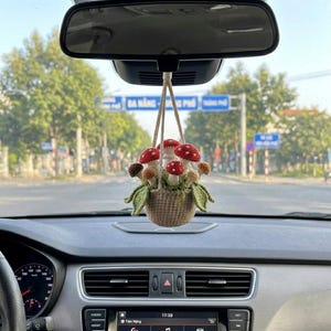 Op de afbeelding: Een handgemaakte auto-hanger met een geweven mandje gevuld met gehaakte paddenstoelen. De paddenstoelen hebben rode hoeden met witte stippen en zijn omgeven door groene bladeren. De hanger hangt aan een touwtje en voegt een vleugje vrolijkheid toe aan het interieur van de auto.
