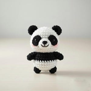 May include: A handmade crochet panda bear with a white body and black accents. The panda has black ears, eyes, and arms, with a white face and a small pink blush on its cheeks. The panda is standing upright.