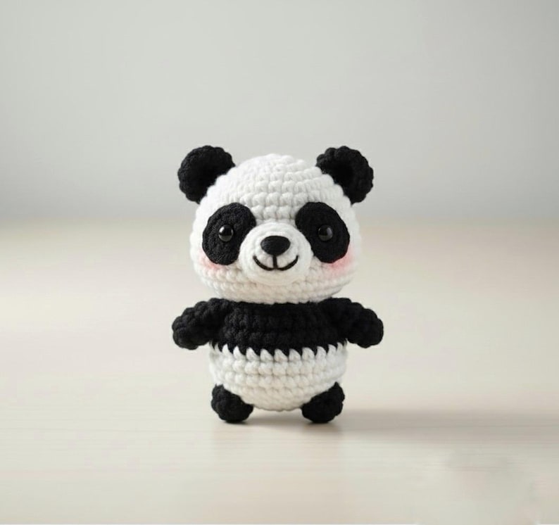 May include: A handmade crochet panda bear with a white body and black accents. The panda has black ears, eyes, and arms, with a white face and a small pink blush on its cheeks. The panda is standing upright.