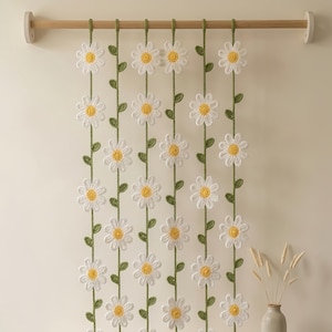 May include: A handmade crochet daisy curtain. White daisy flowers with yellow centers are connected by green stems and leaves. The curtain hangs from a wooden rod, creating a whimsical and decorative home accent.