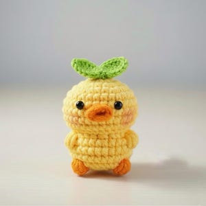 May include: A handmade, yellow crochet chick with an orange beak and feet, and black eyes. The chick has a green leaf detail on top of its head. The crochet work is detailed and the chick has rosy cheeks.
