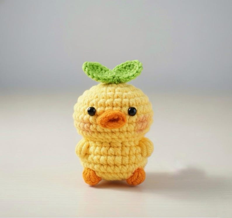 May include: A handmade, yellow crochet chick with an orange beak and feet, and black eyes. The chick has a green leaf detail on top of its head. The crochet work is detailed and the chick has rosy cheeks.