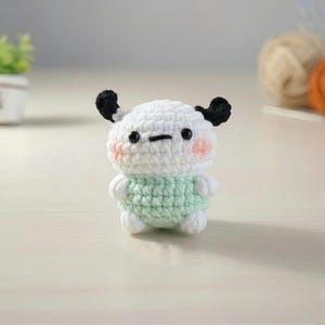 May include: A handmade crochet doll with a white head, black ears, and a light green body. The doll has a simple, cute design with blush-colored cheeks and a small, stitched mouth. The doll is a charming, handcrafted item.