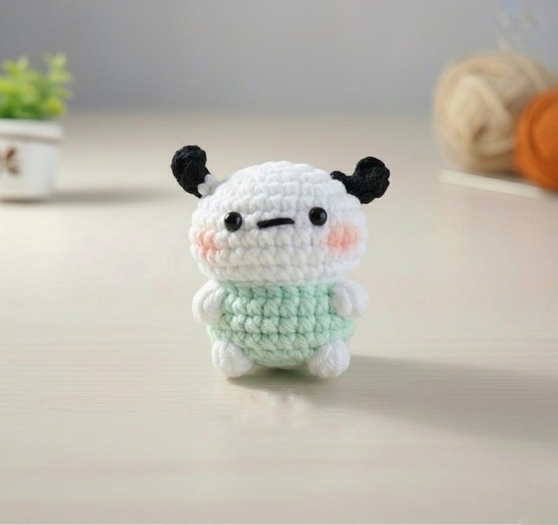 May include: A handmade crochet doll with a white head, black ears, and a light green body. The doll has a simple, cute design with blush-colored cheeks and a small, stitched mouth. The doll is a charming, handcrafted item.