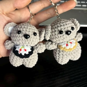 May include: Two handmade crochet koala bear keyrings. The koalas are grey with black eyes and noses. One koala has a black and white bib with red cherries, the other a yellow and white bib with red cherries.
