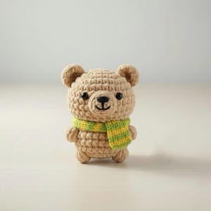 May include: A crocheted teddy bear in tan yarn with black safety eyes and a black nose. The bear wears a green and yellow striped scarf. The handmade bear is a cute, soft toy.