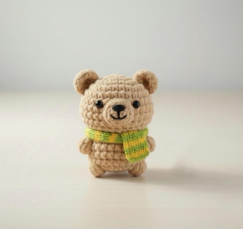 May include: A crocheted teddy bear in tan yarn with black safety eyes and a black nose. The bear wears a green and yellow striped scarf. The handmade bear is a cute, soft toy.