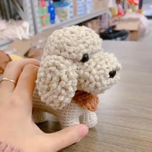Könnte beinhalten: Ein handgefertigter Häkelhund in Creme und Weiß, mit einem strukturierten Körper und einem kleinen braunen Schal. Der Hund hat schwarze Knopfaugen und ein detailliertes, handgefertigtes Aussehen. Das Spielzeug wird von einer Hand gehalten.