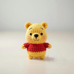 May include: A handmade, yellow and red crochet Winnie the Pooh plush toy. The bear has a yellow head with small ears, a black nose, and a red sweater. The cheeks are pink. The toy is standing on a white surface.