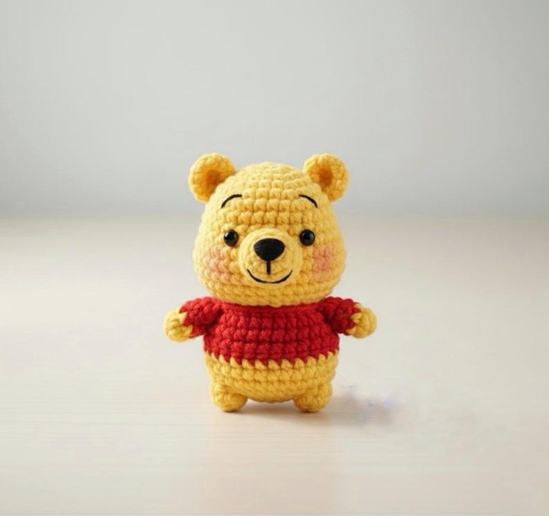 May include: A handmade, yellow and red crochet Winnie the Pooh plush toy. The bear has a yellow head with small ears, a black nose, and a red sweater. The cheeks are pink. The toy is standing on a white surface.
