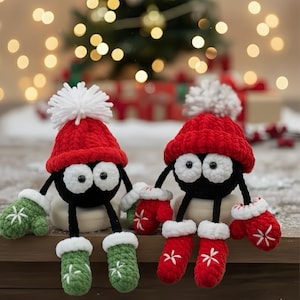 May include: Two handmade Christmas ornaments. Each ornament is a black figure with large white eyes, wearing a red knit hat with a white pom-pom, and matching mittens and boots. One has green mittens and boots with white snowflake embroidery, the other has red mittens and boots with white snowflake embroidery.