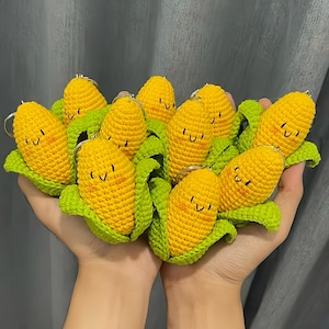 Puede incluir: Una colección de llaveros de maíz hechos a mano con ganchillo. Cada llavero presenta una mazorca de maíz amarilla con una cara sonriente, hojas verdes y un llavero plateado. Los llaveros están diseñados para parecerse a mazorcas de maíz en miniatura.