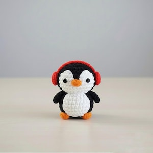 May include: A handmade crochet penguin with a black body, white belly, and orange beak and feet. The penguin wears red headphones. The amigurumi is a cute, small, and soft toy.