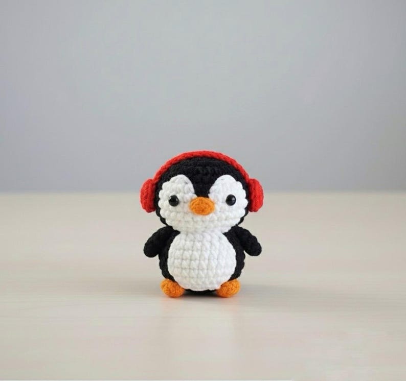 May include: A handmade crochet penguin with a black body, white belly, and orange beak and feet. The penguin wears red headphones. The amigurumi is a cute, small, and soft toy.