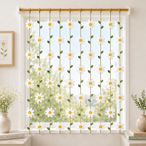 May include: A window curtain featuring ten vertical green crocheted strands adorned with white daisy flowers with yellow centers. The strands are suspended from a horizontal wooden rod, creating a floral pattern across the window frame.