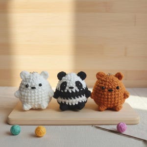 May include: Three crocheted bear-shaped keychains in white, black and white panda, and orange. They have small black eyes and are standing on a wooden board. Colorful yarn balls and a crochet hook are also present.