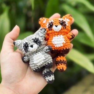 Puede incluir: Dos pequeñas figuras de animales de ganchillo hechas a mano. Una es un mapache gris y blanco, la otra un panda rojo naranja y marrón. Ambos tienen ojos negros y están sostenidos en la mano de una persona. El fondo es follaje verde borroso.