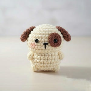 May include: A crocheted dog figurine in cream, brown, and black. The handmade amigurumi dog has brown ears, a brown spot over one eye, and a black nose and eyes. The dog has a blush pink cheek.