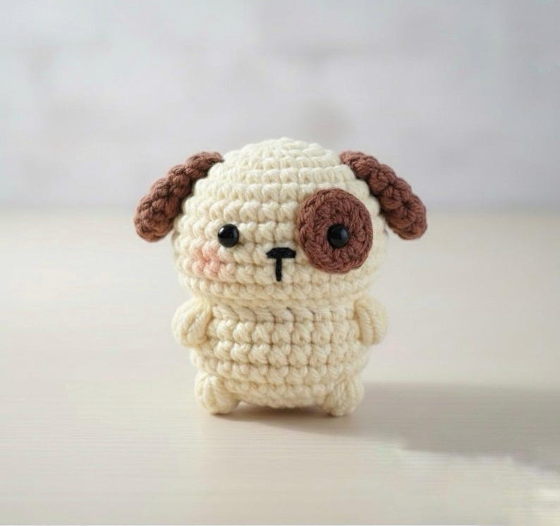 May include: A crocheted dog figurine in cream, brown, and black. The handmade amigurumi dog has brown ears, a brown spot over one eye, and a black nose and eyes. The dog has a blush pink cheek.