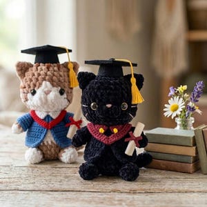 Puede incluir: Dos figuras de gato de ganchillo, una marrón y blanca, la otra negra, ambas con birretes de graduación con borlas amarillas y diplomas en miniatura. El gato marrón lleva una chaqueta azul con ribete rojo, el gato negro un cuello rojo y dorado.