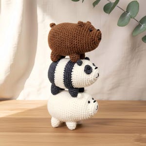 May include: Three crocheted bear figurines stacked on top of each other. The top bear is brown, the middle bear is a panda with black and white stripes, and the bottom bear is white. The bears are on a wooden surface.