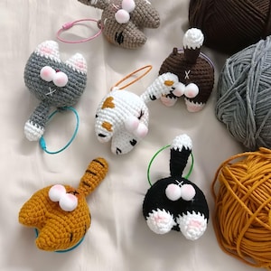 Op de afbeelding: Een verzameling handgemaakte gehaakte kattenkont haarbanden in verschillende kleuren, waaronder grijs, wit, bruin, zwart en oranje. Elke kattenkont heeft een pluizige staart en roze accenten. Garenbollen zijn zichtbaar op de achtergrond.