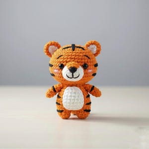 May include: A handmade crochet tiger toy in orange, black, and white. The tiger has a white belly, black stripes, and a smiling face with pink cheeks. The toy is standing upright.