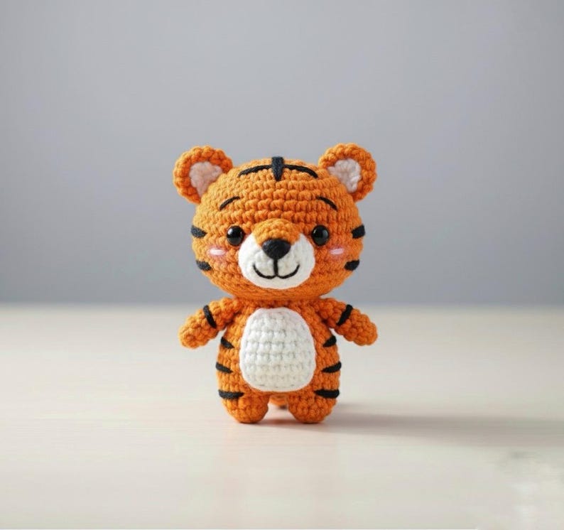 May include: A handmade crochet tiger toy in orange, black, and white. The tiger has a white belly, black stripes, and a smiling face with pink cheeks. The toy is standing upright.