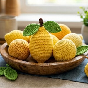 Puede incluir: Un cuenco de madera lleno de varios limones de ganchillo y un solo limón con hojas verdes. Los limones son de varios tonos de amarillo y naranja. Las hojas son verdes. El cuenco está sobre una tela azul.