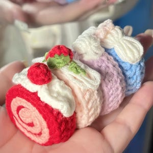 Puede incluir: Colección de cuatro pasteles de ganchillo en miniatura. El primero es un brazo de gitano rojo y blanco con una fresa encima. Los otros tres son de colores pastel, parecidos a pequeños pasteles con glaseado blanco. Los pasteles se sostienen en una mano.