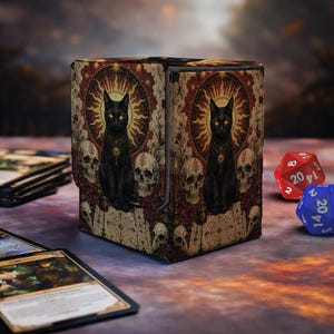 Puede incluir: Una caja para cartas rectangular con una ilustración oscura de un gato negro frente a un sol dorado, rodeado de calaveras humanas y patrones geométricos complejos en tonos rojo, dorado y negro, junto a dos dados de veinte caras.