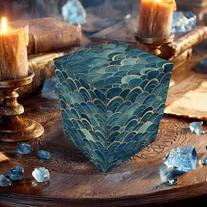 May include: A teal and gold art deco patterned cube sits on a wooden table, surrounded by candles, books, and crystals. The cube's design features a repeating fan-like pattern. Candles are lit, creating a warm ambiance.