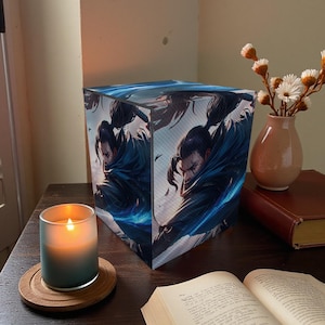 May include: A cube-shaped lamp featuring a digital art print of a warrior in a blue cloak. A lit candle in a frosted glass and a small vase with white flowers are on a wooden table. An open book and a coaster are also visible.