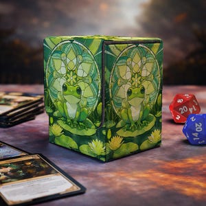May include: A square deck box for trading cards featuring a green frog sitting on a lily pad. The design includes a stained glass style circular pattern with lotus flowers in shades of green and yellow. The box has a textured fabric finish.