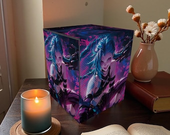 May include: A cube-shaped lamp featuring a vibrant illustration of a character with blue hair and a mischievous expression. The artwork is set against a backdrop of purple and pink hues. A lit candle and a vase of flowers are also visible.