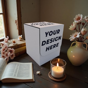 May include: A white box with the text "YOUR DESIGN HERE" in navy blue. The box is on a dark wooden surface, next to a lit candle in a glass holder, a vase of flowers, and an open book.