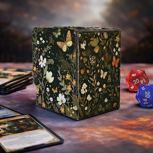 May include: A dark green card storage box with a floral and butterfly pattern. The box is decorated with white flowers, orange and yellow butterflies, and green foliage. Two dice and a few cards are visible in the image.