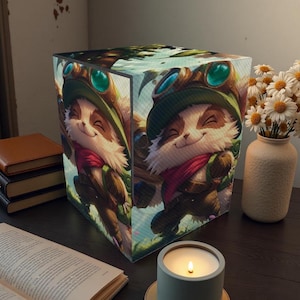 Puede incluir: Lámpara cúbica con un personaje de dibujos animados con sombrero verde, gafas y bufanda roja. La lámpara está sobre una superficie oscura junto a una vela encendida, un jarrón con flores y libros.